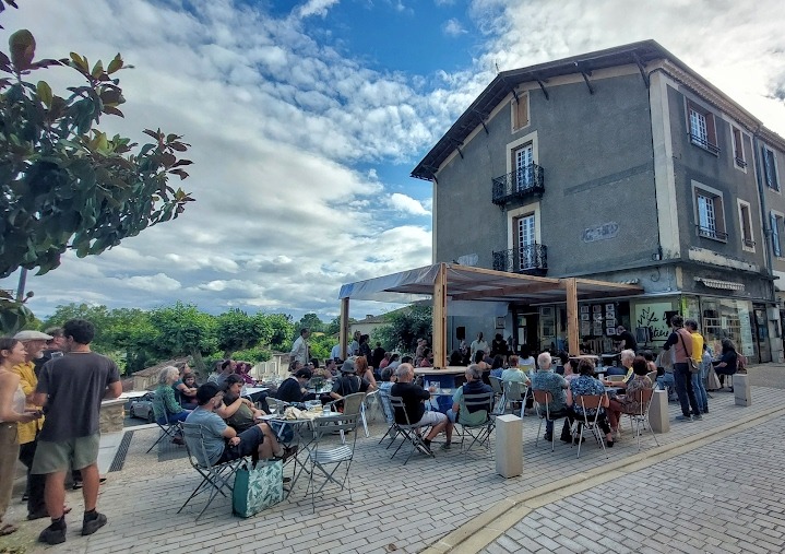 Terrasse de café animée avec clients assis et debout devant bâtiment
